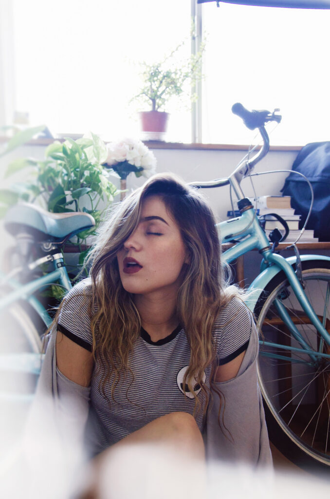Darian Rojas a stylish individual with wavy hair is seated indoors near a window. She wears a striped shirt with a unique emblem and has a relaxed demeanor. The room is adorned with plants books and a bicycle giving it a cozy and artistic vibe.